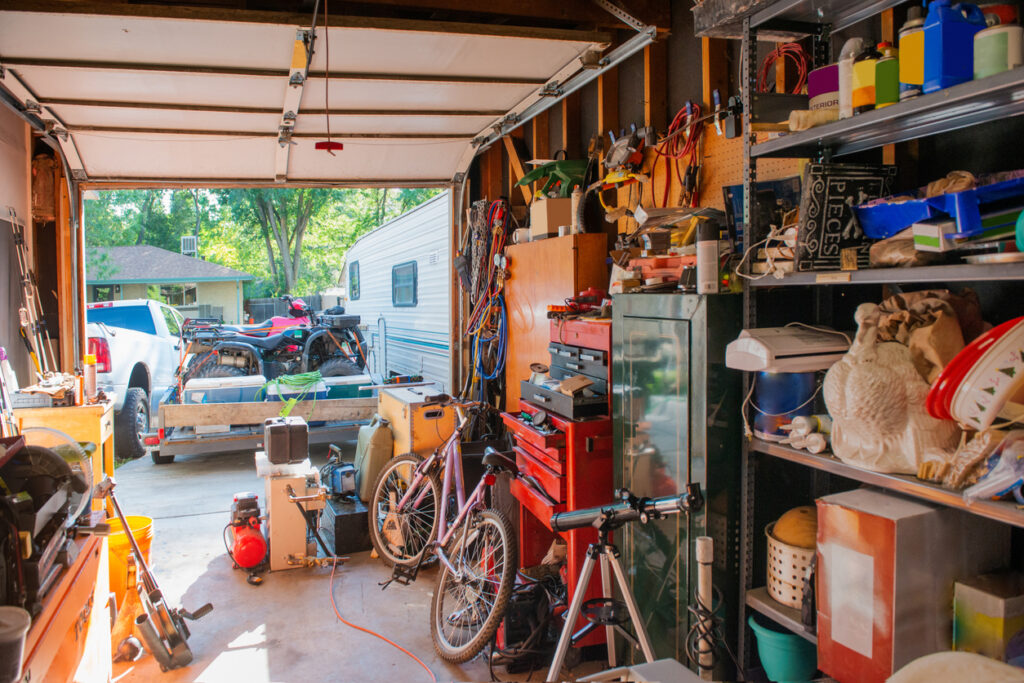 A garage lined with shelves full of things stored at home including, tools, cleaning supplies, holiday decorations, and sporting equipment. The garage door is open.