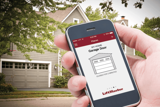 A residential home sits in the background. In the foreground, there is a close-up of a hand holding a smartphone. The screen shows a LiftMaster app, showcasing how you can control your garage door from your phone.