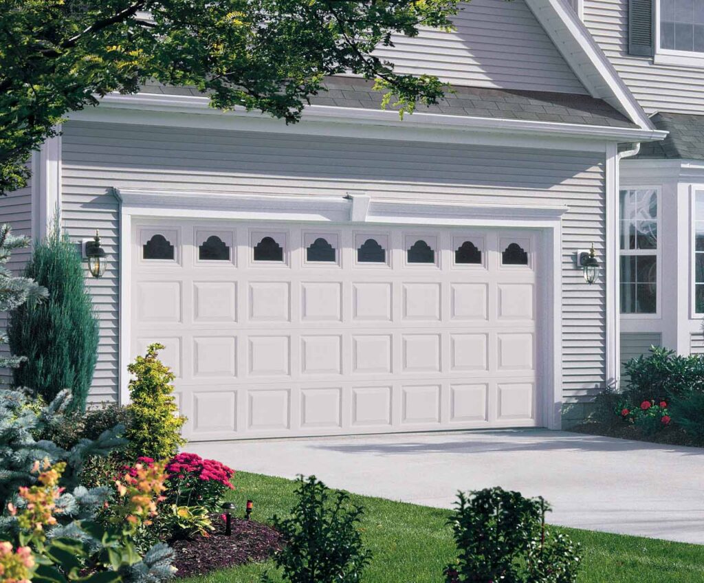 White paneled garage door with small windows across the top, in a large two-story white home with trees and landscaping to the left.